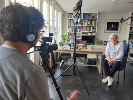 Een oudere vrouw met wit haar zit in een zetel, ze wordt geïnterviewd voor een documentaire over het dichten van de loonkloof